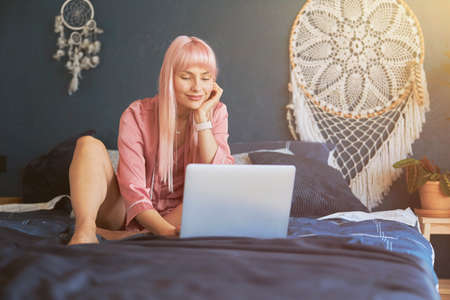 Lady with long dyed pink hair surfs internet with laptop resting on bedの写真素材