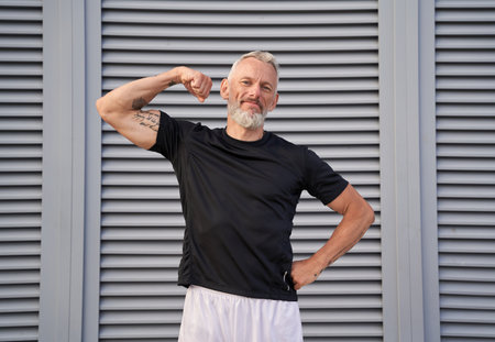 Sporty mature man looking at camera, showing his muscular arm and bicep while standing outdoors over gray backgroundの写真素材