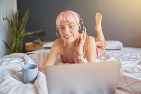 Cheerful woman with headphones and cup rests using laptop on comfortable bedの写真素材