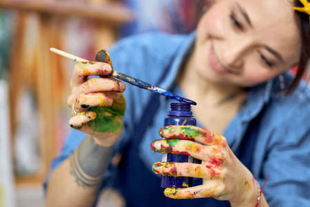 Smiling young woman in apron holding blue paint jar and a paintbrush, working on a painting in workshopの写真素材