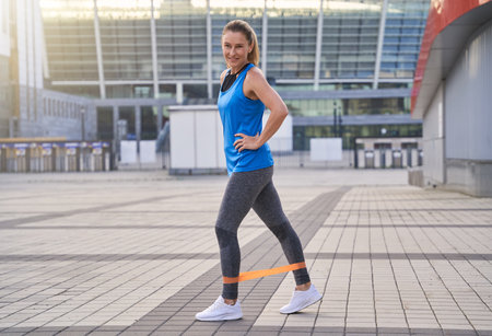 Full length shot of happy sporty woman in sportswear warming up using resistance band while standing outdoors, exercising in the cityの写真素材