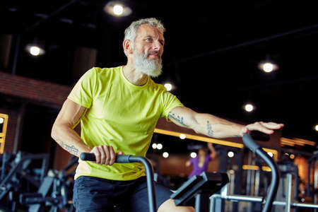 Exercises after 50. Side view of a mature athletic man in sportswear doing cycling on exercise bikes at gymの写真素材