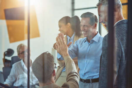 Two excited coworkers giving high five to each other and smiling while having a meeting with colleagues in the modern officeの写真素材