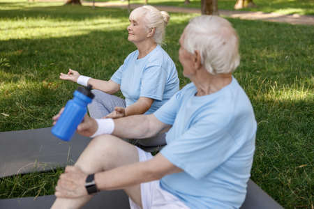 Tranquil senior lady meditates sitting near man with bottle of water on field grassの写真素材
