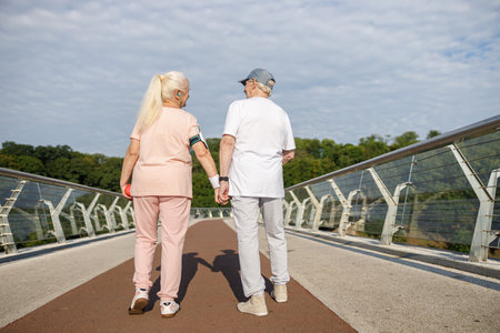 Positive senior woman and man walk holding hands along footbridge after trainingの写真素材