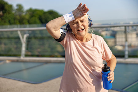 Joyful mature woman with smartphone in headphones mops brows at training on bridgeの写真素材