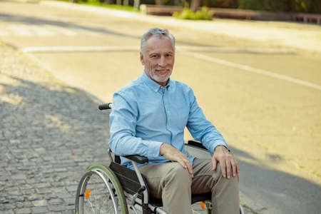 Elderly man in wheelchair smiling at camera while riding alone in the park on a summer dayの写真素材