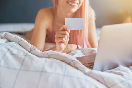 Smiling woman with holds blank credit card near laptop during online shopping o bedの写真素材