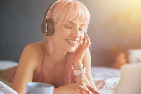 Lady with pink hair and headphones uses modern laptop resting on bedの写真素材