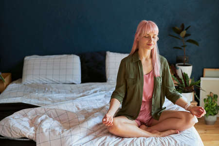 Positive young woman in green shirt and pajamas meditates sitting in lotus pose on large bedの写真素材