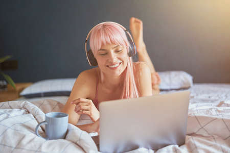 Positive woman with headphones and cup watches video via laptop on bedの写真素材