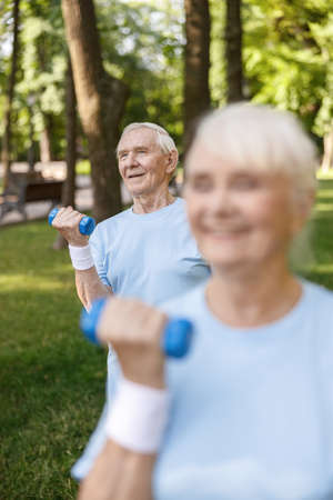Happy senior gentleman does exercises with dumbbell training with wife in picturesque parkの写真素材