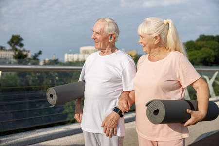 Cheerful senior couple with mats listens to music standing on footbridgeの写真素材