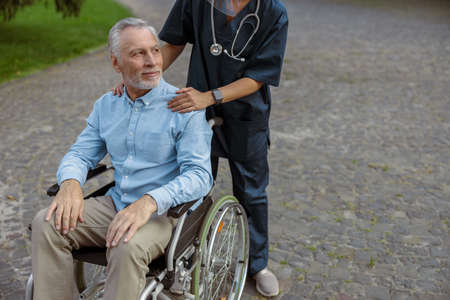 Recovering male patient in a wheelchair looking away on a walk with a nurse outdoorsの写真素材