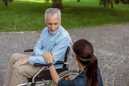 Senior aged man, recovering patient in wheelchair looking and holding hand of nurse wearing face shield and mask, supporting himの写真素材