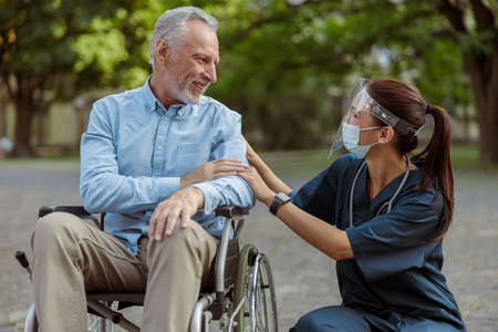 Grateful mature man, recovering patient in wheelchair having conversation with a nurse wearing face shield and mask on a walk in the parkの写真素材