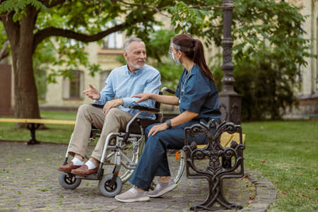 Full length shot of cheerful mature man, recovering patient in wheelchair talking to caring nurse, resting in the park near hospitalの写真素材
