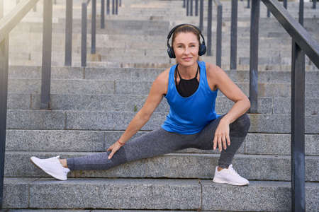 Fit middle aged woman in headphones looking at camera while stretching legs, having workout outdoors in the cityの写真素材