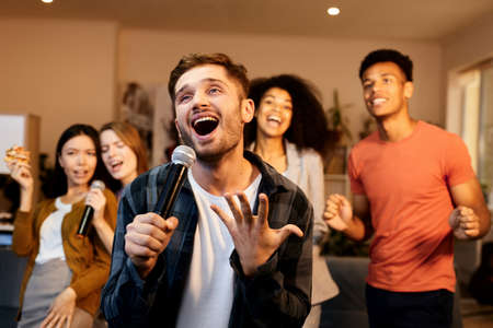 Fun at home. Enthusiastic handsome young guy singing with microphone while playing karaoke with friends indoors, standing in the modern apartmentの写真素材
