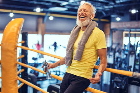 Happy mature man with towel on shoulders looking at camera and smiling, standing on the ring and resting after boxing training at gymの写真素材