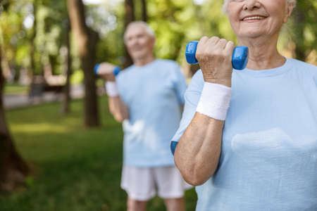 Sportive senior woman with sweatband does exercises with dumbbell training with friend in parkの写真素材