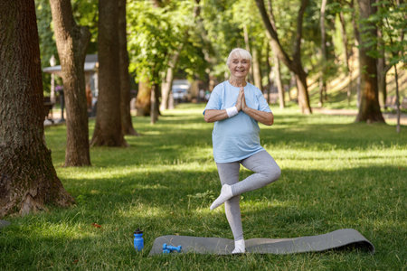 Happy senior lady in tracksuit does tree pose practicing yoga asana in parkの写真素材