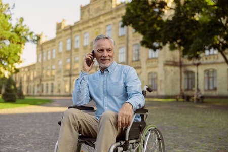Smiling aged man recovering patient in wheelchair looking away while talking on the phone, spending time alone in the park near hospitalの写真素材