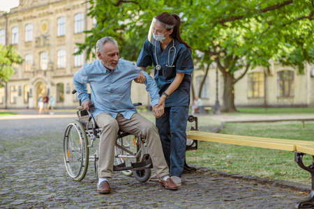 Full length shot of caring nurse wearing face shield and mask helping mature man, recovering patient in wheelchair to stand up in the park near hospitalの写真素材