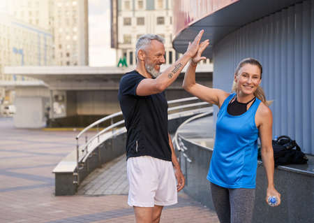 Joyful sportive middle aged friends, man and woman in sportswear giving high five, standing together outdoors ready for workoutの写真素材