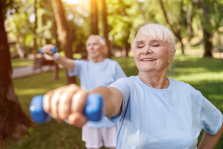 Strong senior woman does exercises with dumbbell training with husband in green parkの写真素材