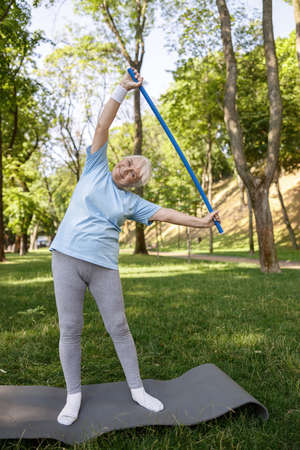 Senior lady in tracksuit does side bend with plastic bar standing in green parkの写真素材
