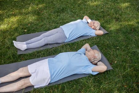 Lovely senior family couple looks at each other lying on mats after training in green parkの写真素材