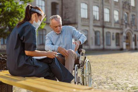 Recovering male patient in wheelchair discussing his health condition with nurse in protective face shield outdoors near rehabilitation clinicの写真素材