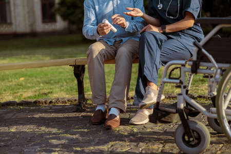 Cropped shot of elderly recovering male patient having conversation with a nurse, sitting together on the bench near rehabilitation clinicの写真素材