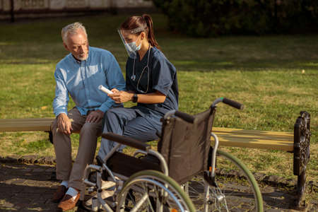 Serious aged recovering male patient listening to nurse wearing face shield, sitting together on the bench near rehabilitation clinicの写真素材