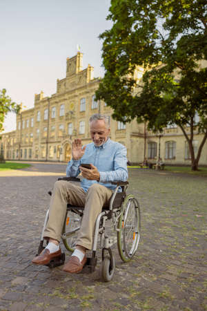 Smiling senior man, recovering male patient in wheelchair making a video call using smartphone while resting alone in the park near hospitalの写真素材