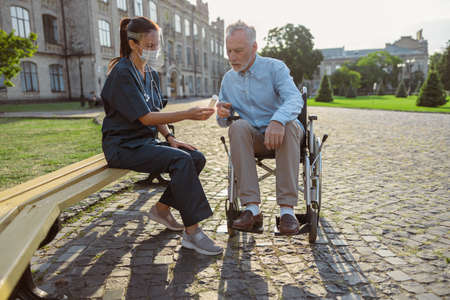 Focused senior man recovering patient listening to prescription of nurse wearing face shield, resting together outdoors near rehabilitation clinicの写真素材
