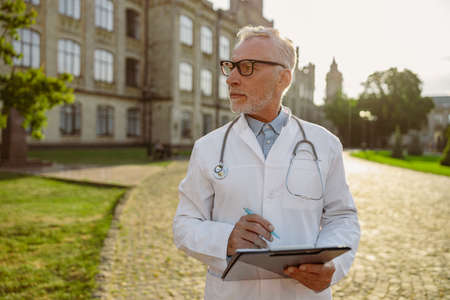 Serious senior male doctor in lab coat and glasses holding clipboard, looking aside, standing outdoors near clinicの写真素材
