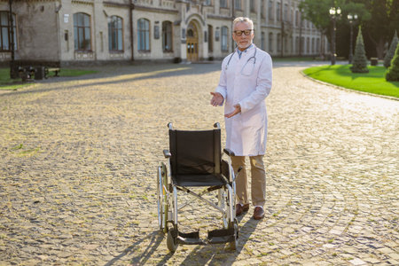 Full length shot of senior male doctor in lab coat and glasses looking at camera, welcoming patient, standing near wheelchair outdoorsの写真素材