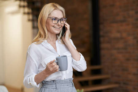 Morning. Cheerful middle aged woman with blonde hair and glasses holding a cup of coffee or tea while talking on her phone, standing at homeの写真素材