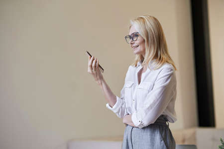 Busy hours. Stylish mature blonde woman in glasses and business attire smiling and talking on the phone, standing indoorsの写真素材