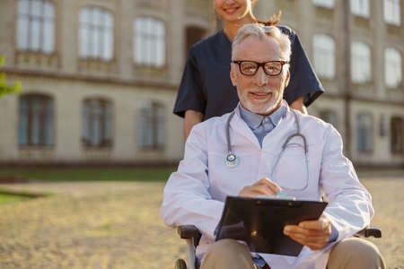 Busy senior handicapped male doctor in wheelchair wearing lab coat and glasses with young nurse assisting him outdoorsの写真素材