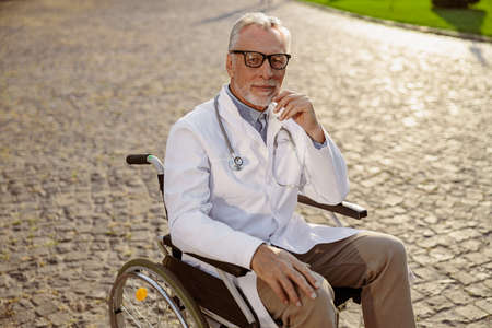 Pensive senior handicapped male doctor in wheelchair wearing lab coat and glasses looking at camera, posing outdoors on a sunny dayの写真素材