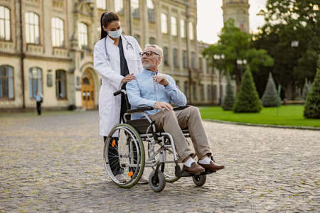 Full length shot of senior man handicapped patient in wheelchair smiling at young nurse in protective mask helping him outdoorsの写真素材