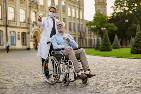 Full length shot of caring young nurse in protective mask taking care of senior man handicapped patient in wheelchair outdoorsの写真素材