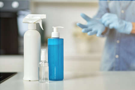 Sanitizer bottles to clean surfaces in the kitchen. Woman putting on protective gloves while getting ready for clean up in the background. Antiseptic, disinfection, cleanliness and healthcareの写真素材