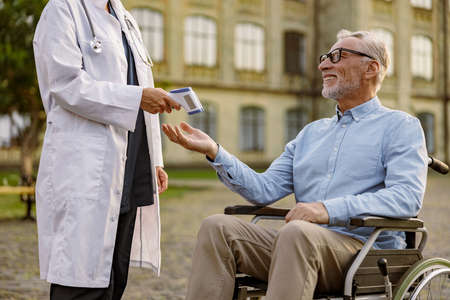 Smiling senior handicapped man reaching out hand for doctor to check fever by digital thermometer. Scanning and protection from coronavirusの写真素材