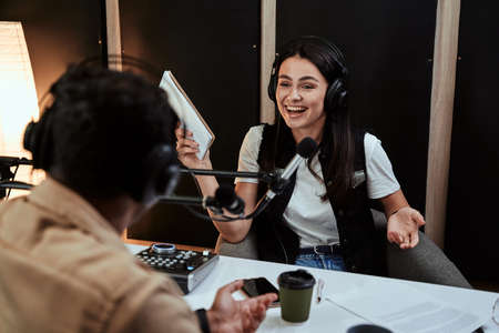 Portait of happy female radio host laughing, listening to male guest, presenter and holding a script paper while moderating a live show in studioの写真素材