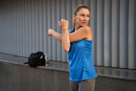 Sportive mature woman in sportswear looking away while warming up, stretching her arms, standing outdoorsの写真素材