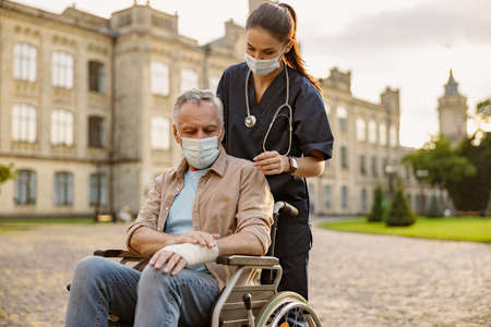 Young nurse in protective mask taking care of senior recovering patient in wheelchair with bandaged hand outdoors in the park near clinicの写真素材